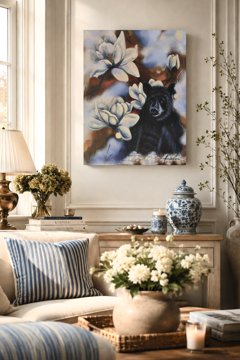 Original painting "Feeling Small in a Big World" by Annikaprana, displayed above a sideboard in a bright classic living room with cream tones and blue accents — a moody oil painting of a black bear surrounded by large white magnolia blossoms