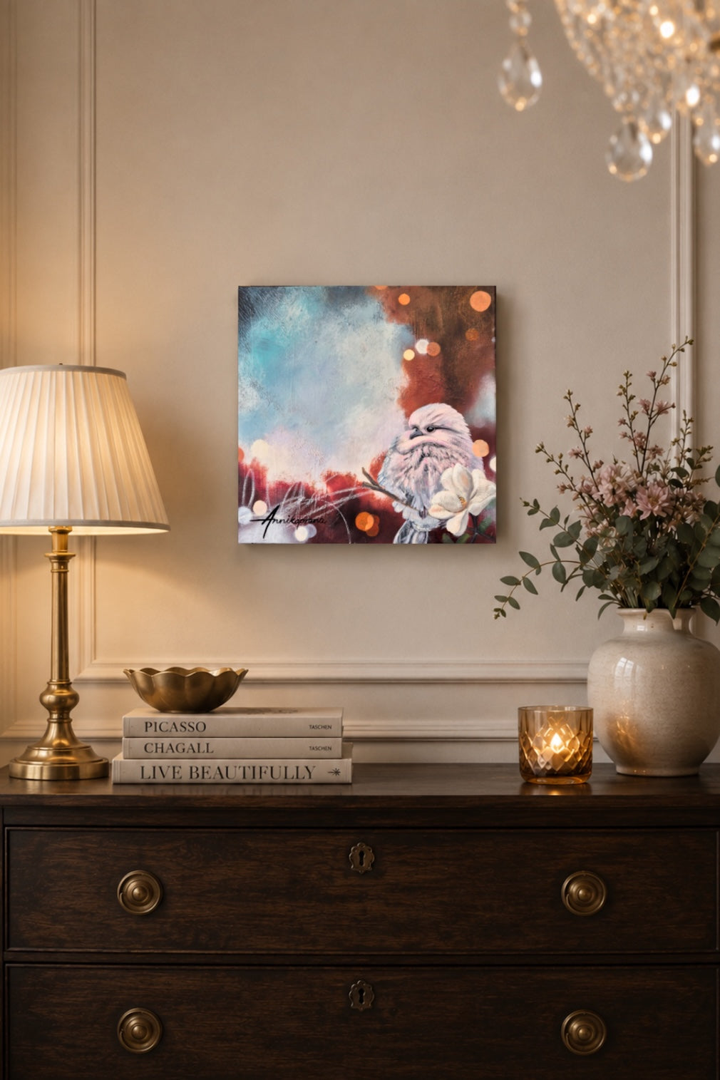 "Free as a Bird" by Annika Andersson — original painting displayed on a wall above a dark wooden dresser in an elegant interior with warm lamplight, art books, and a white vase with branches