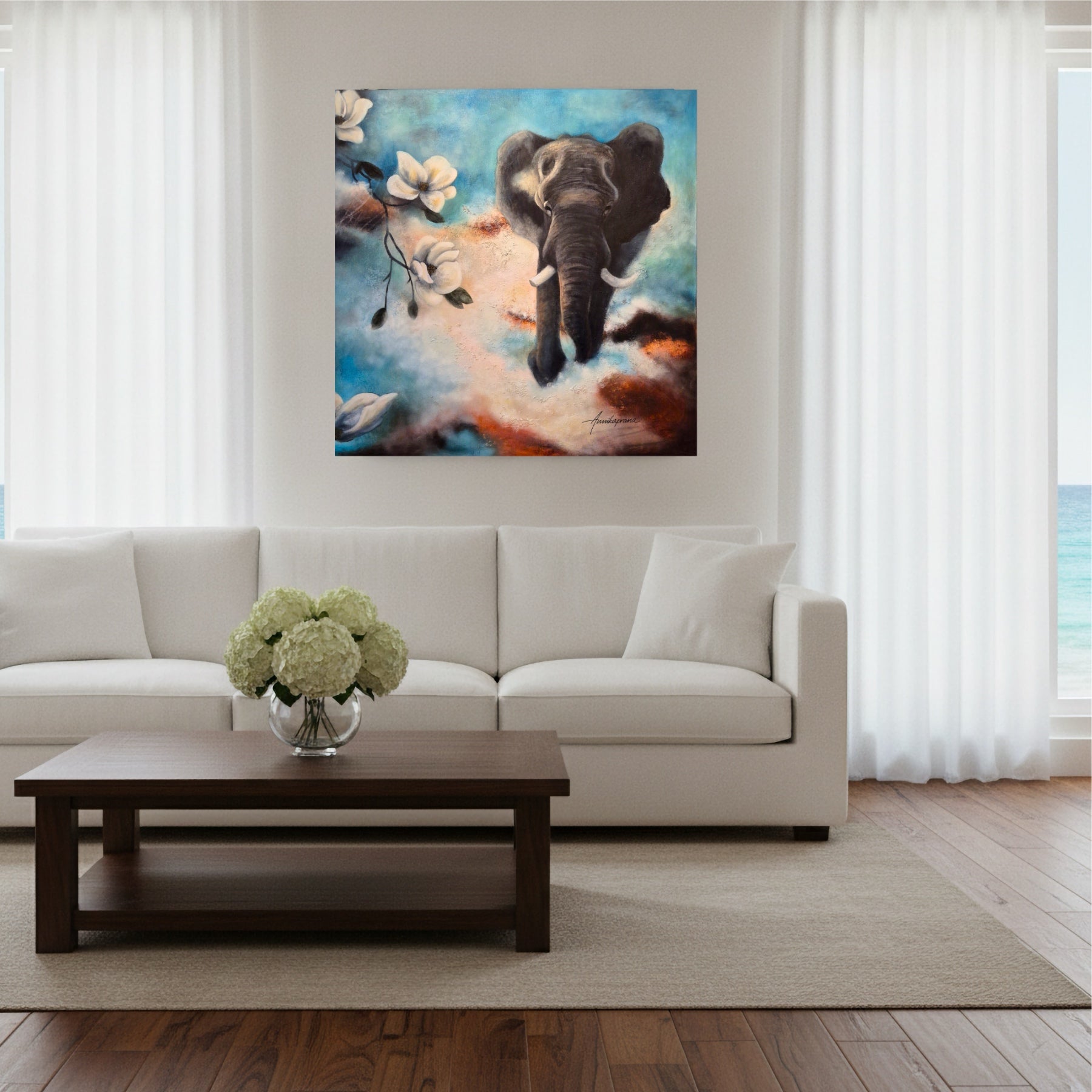 Canvas print "The Power of Stepping into the Light" by Annikaprana, displayed above a white sofa in a bright coastal living room — a dramatic painting of an elephant emerging through clouds and white magnolia blossoms in shades of blue, terracotta and cream