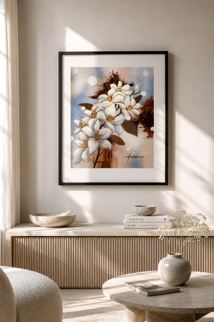 "Snow White Lilies 2025 — original painting by Annikaprana in a black wood frame with white passepartout in a warm Scandinavian living room, white lilies with golden centres and warm brown tones on a soft blue-grey background, Mixed Media on Fabriano paper, signed"