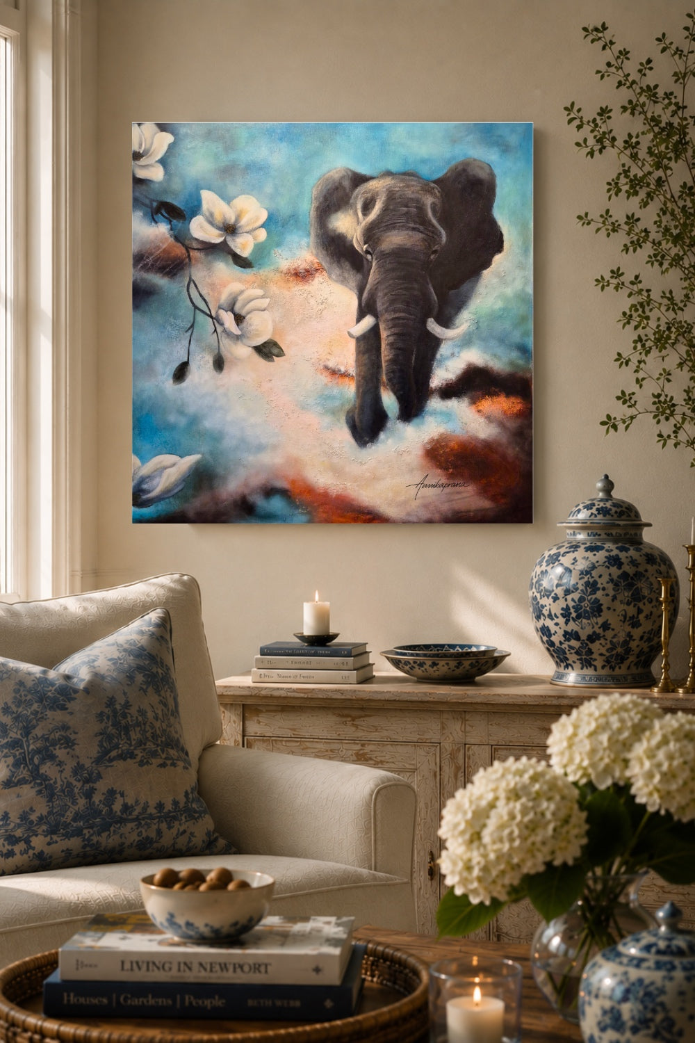 Canvas print "The Power of Stepping into the Light" by Annikaprana, displayed above a sideboard in a warm classic living room with cream tones and blue accents — a dramatic painting of an elephant emerging through clouds and white magnolia blossoms in shades of blue, terracotta and cream