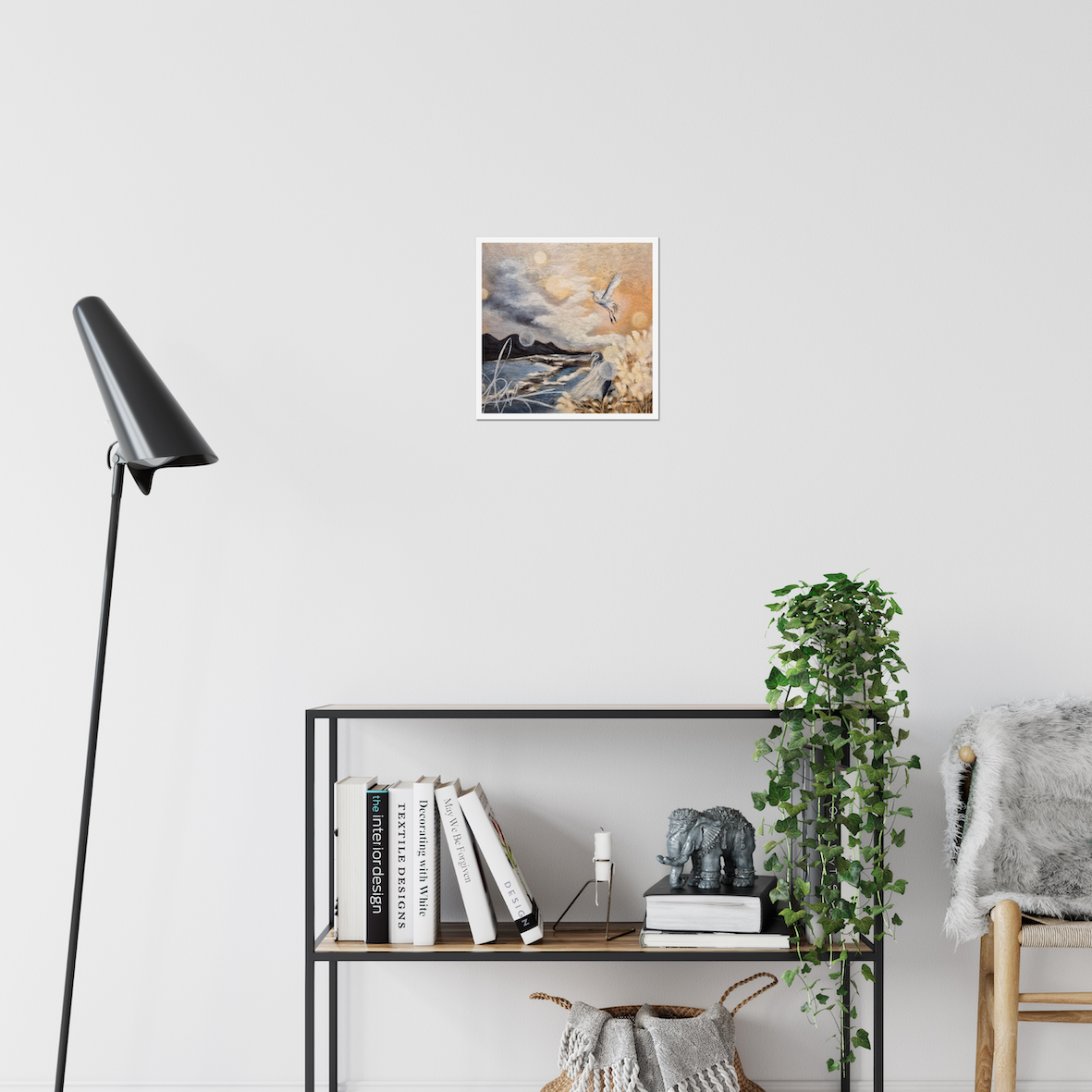 New Horizons Ahead — Fine Art Print by Annikaprana. A white bird soaring above a dramatic coastal landscape in warm amber and stormy grey tones. Giclée on Hahnemühle German Etching 310gsm, white border, 12×12″ (≈30×30 cm). Displayed on a white wall in a Scandinavian living room with a black floor lamp and open bookshelf.