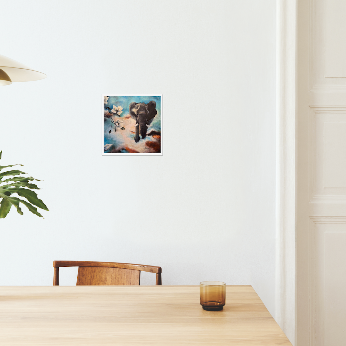 The Power of Stepping into the Light — Fine Art Print by Annikaprana. A majestic elephant emerging from swirling clouds among white magnolia blossoms in turquoise and warm amber tones. Giclée on Hahnemühle German Etching 310gsm, white border, 12×12″ (≈30×30 cm). Displayed on a white wall in a Scandinavian dining room.